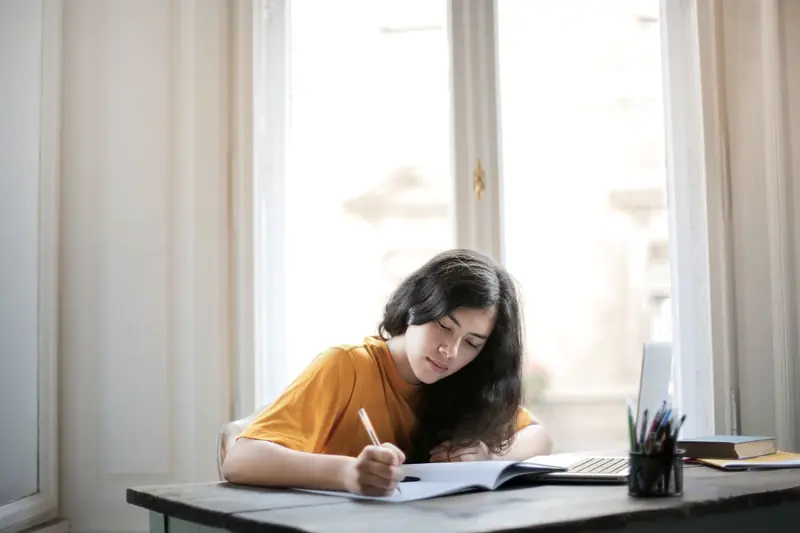 Una joven escribe en un cuaderno junto a una ventana, con la computadora al lado