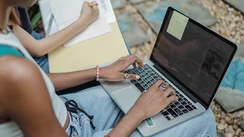 Manos de dos jóvenes tomando apuntes y escribiendo en una computadora portátil